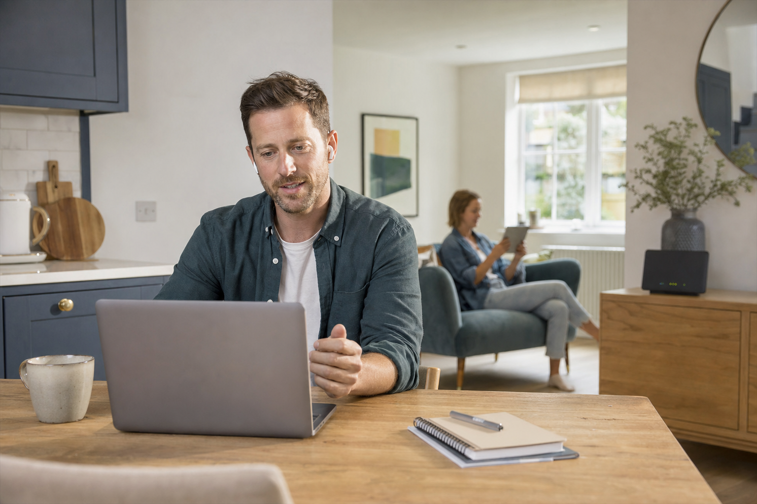 Clean modern UK living room in natural daylight with a person on a calm video call on a laptop at a kitchen table, another person in the background using a tablet, and a discreet broadband router on a sideboard, suggesting steady video calls over home broadband